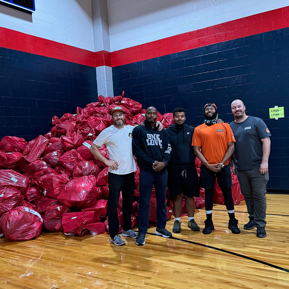 A team of movers standing in a gymnasium in front of a large pile of bagged donations.