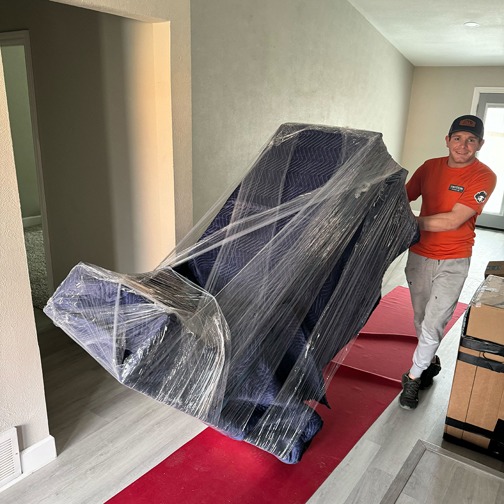 A mover pushing a wrapped piece of furniture down a hallway lined with a red carpet to protect the floor.