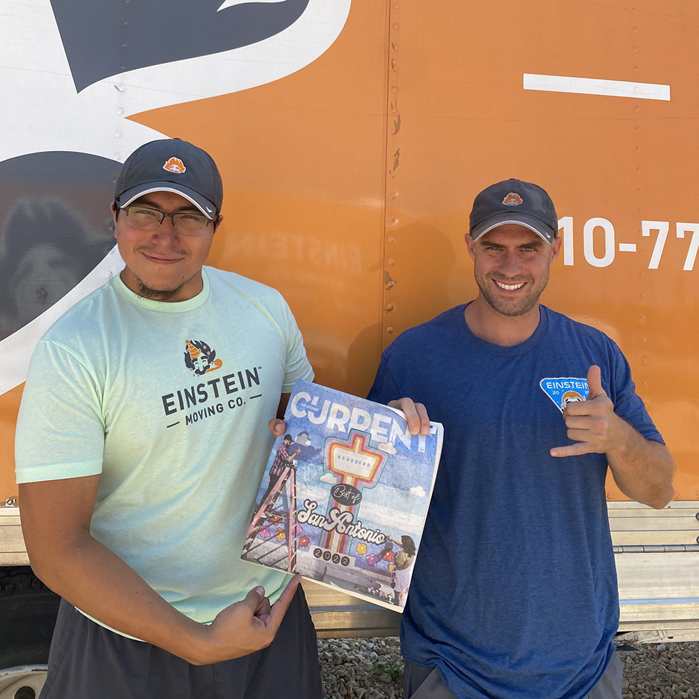 Two Einstein Moving Company team members smiling and holding a magazine cover that says 'Best of San Antonio,' standing in front of an orange company truck.