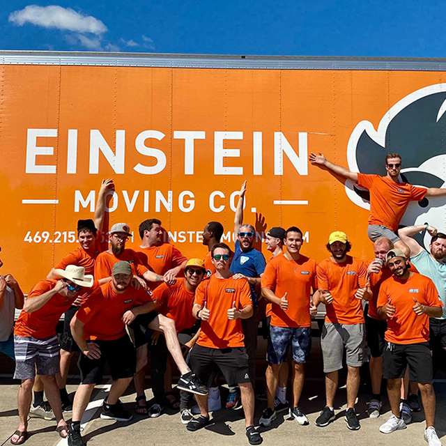 Group of Einstein Moving Company team members in orange shirts posing energetically in front of a branded moving truck.